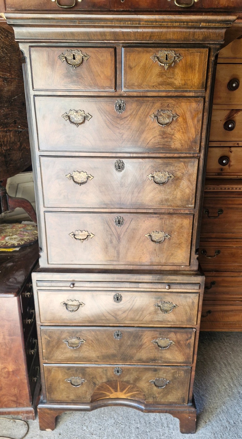 An early 20th century walnut chest on chest featuring brass drop handles, a pull-out brushing slide, and an inlaid sunburst motif on the lower drawer. View VII