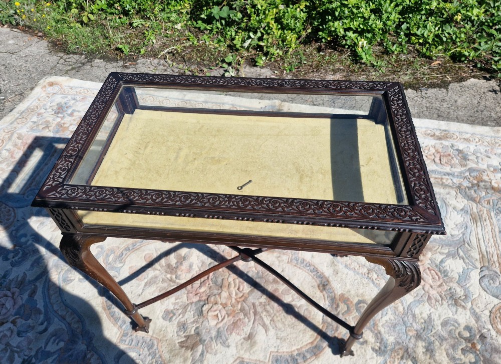 An Edwardian carved mahogany bijouterie table with glass display panels, cabriole legs, and an X-frame stretcher.