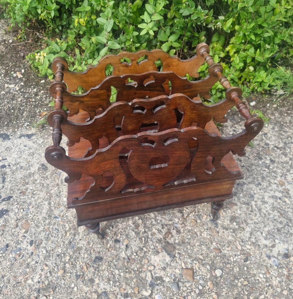 A Victorian carved rosewood Canterbury featuring four open partitions with scrollwork dividers, a single drawer with turned knobs, and turned legs on brass castors. View III