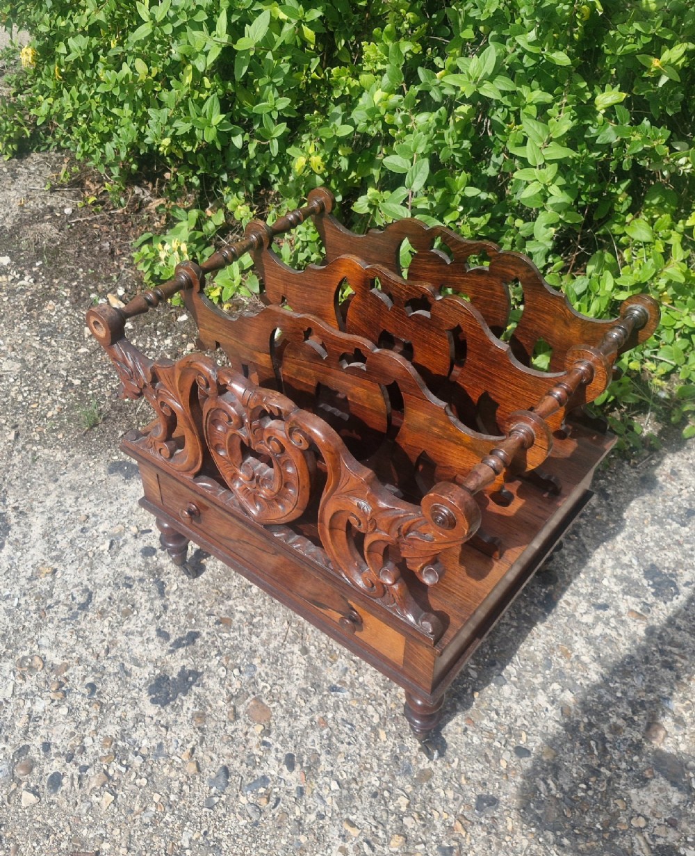 A Victorian carved rosewood Canterbury featuring four open partitions with scrollwork dividers, a single drawer with turned knobs, and turned legs on brass castors. View IV
