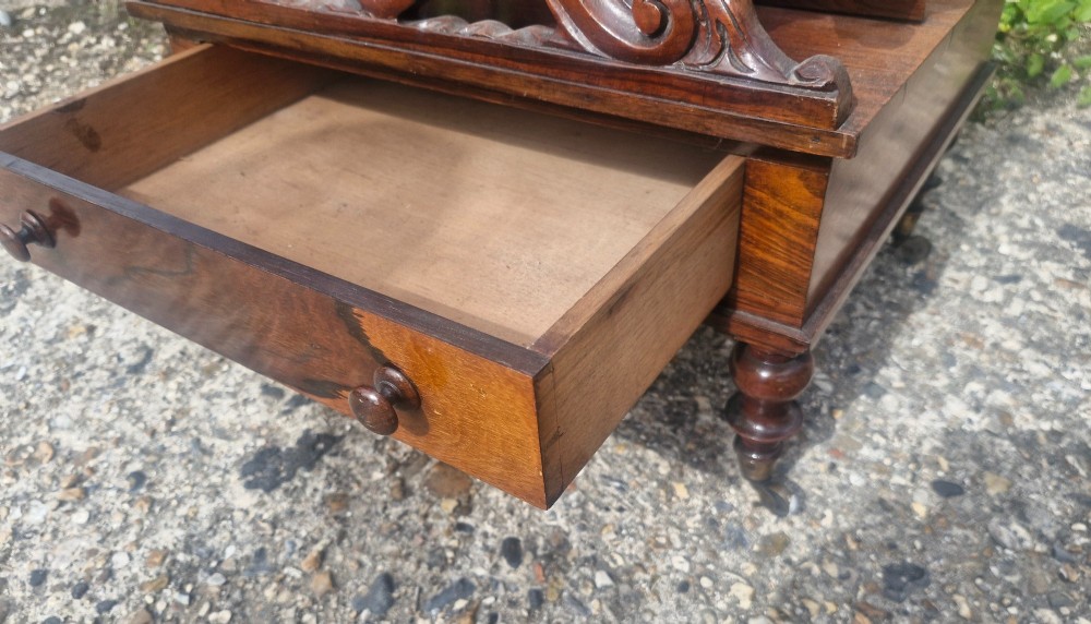 A Victorian carved rosewood Canterbury featuring four open partitions with scrollwork dividers, a single drawer with turned knobs, and turned legs on brass castors. View V