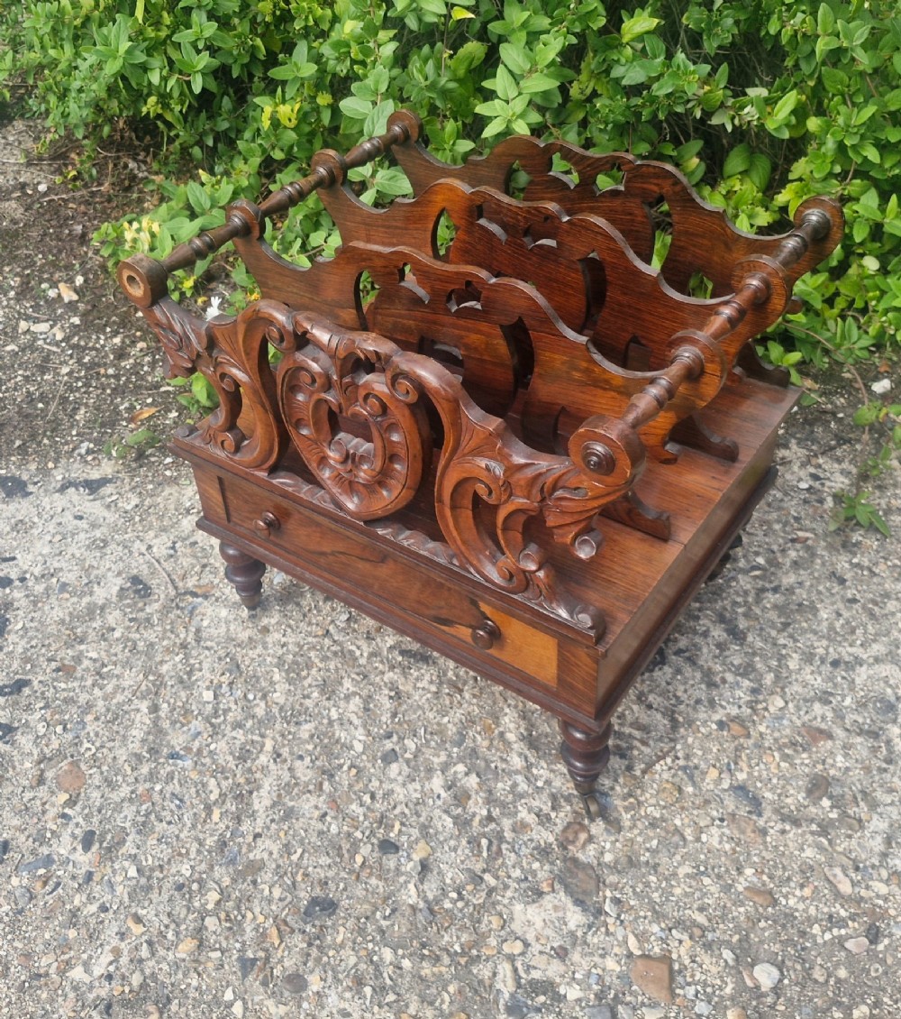 A Victorian carved rosewood Canterbury featuring four open partitions with scrollwork dividers, a single drawer with turned knobs, and turned legs on brass castors. View VI