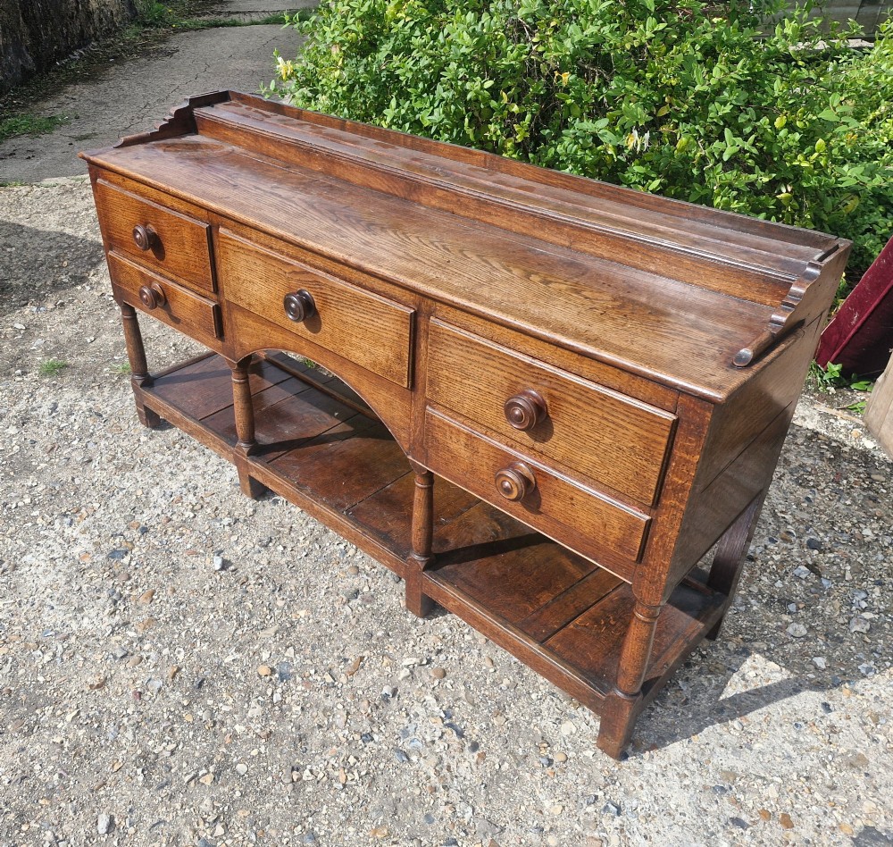A Regency two-part Welsh oak dresser featuring an upper open three-shelf plate rack above five drawers, an arched central apron, turned supports, and a lower pot board shelf. View II