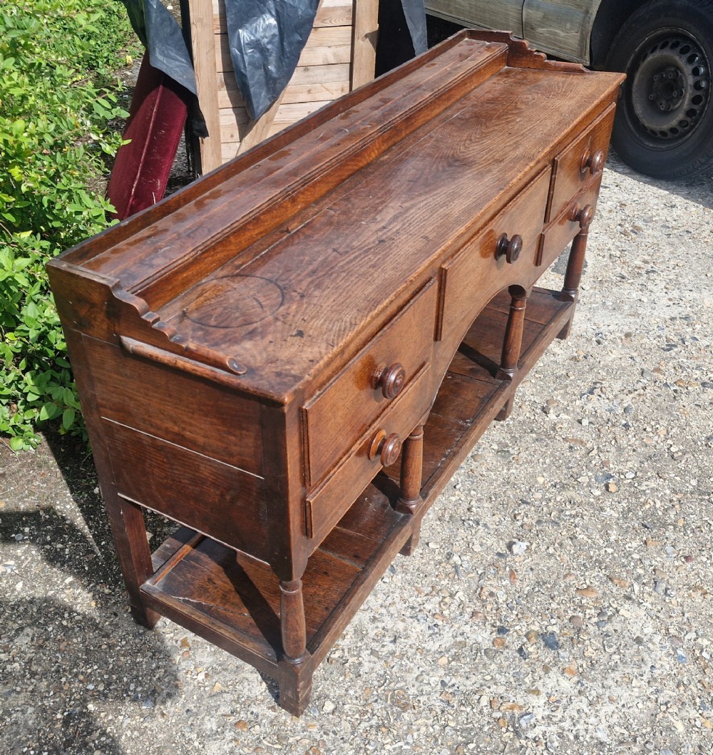 A Regency two-part Welsh oak dresser featuring an upper open three-shelf plate rack above five drawers, an arched central apron, turned supports, and a lower pot board shelf. View V