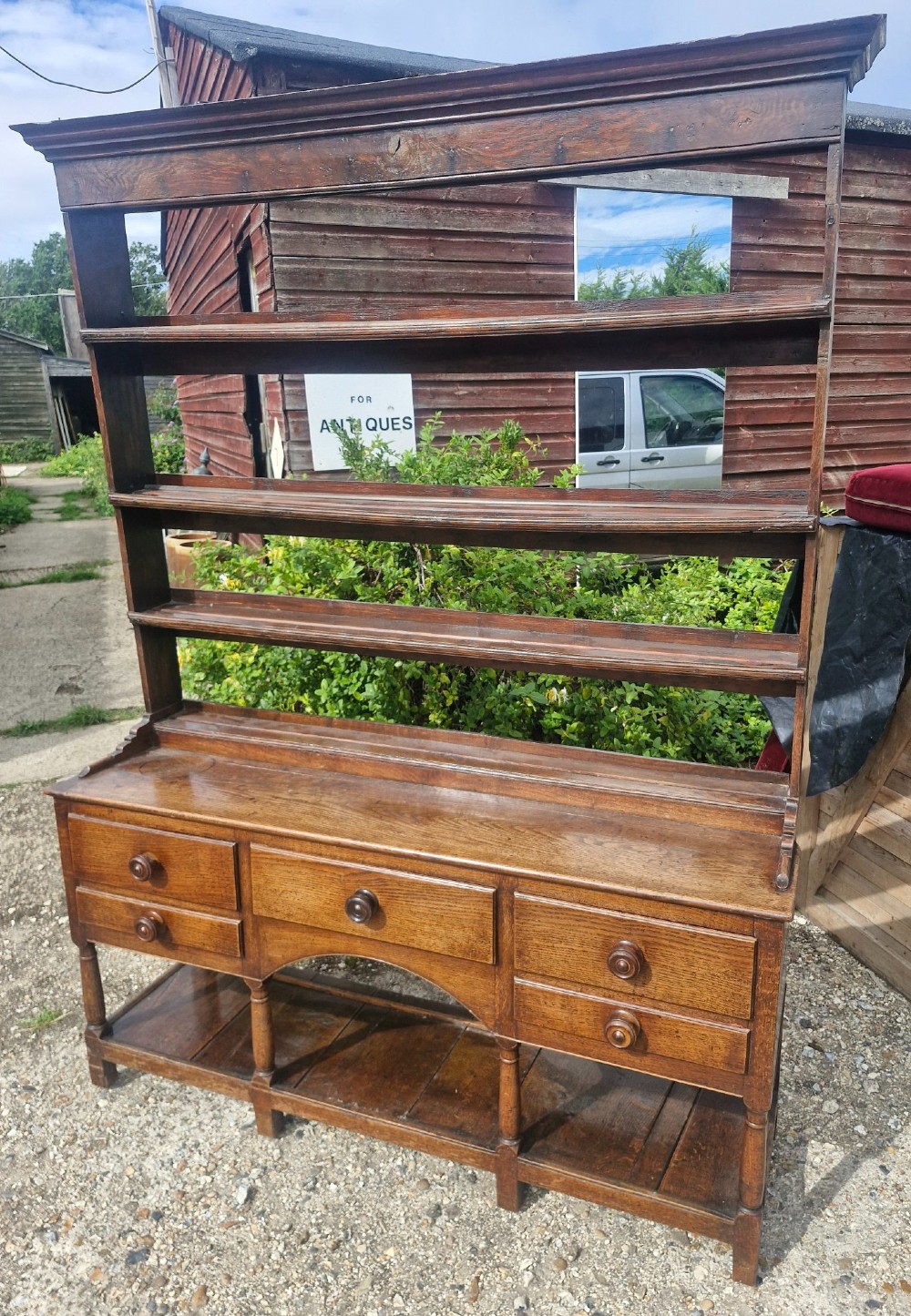 A Regency two-part Welsh oak dresser featuring an upper open three-shelf plate rack above five drawers, an arched central apron, turned supports, and a lower pot board shelf. View VII