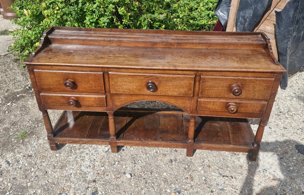 A Regency two-part Welsh oak dresser featuring an upper open three-shelf plate rack above five drawers, an arched central apron, turned supports, and a lower pot board shelf. View VIII