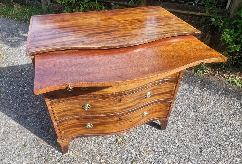 An antique mahogany chest of drawers featuring a serpentine curved front, four graduated drawers with circular brass pull handles, a pull-out brushing slide, and inlaid stringing detail. View II