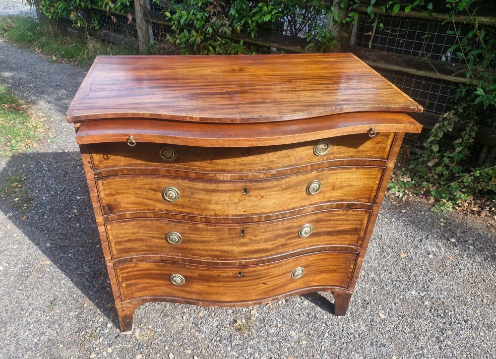 An antique mahogany chest of drawers featuring a serpentine curved front, four graduated drawers with circular brass pull handles, a pull-out brushing slide, and inlaid stringing detail. View III
