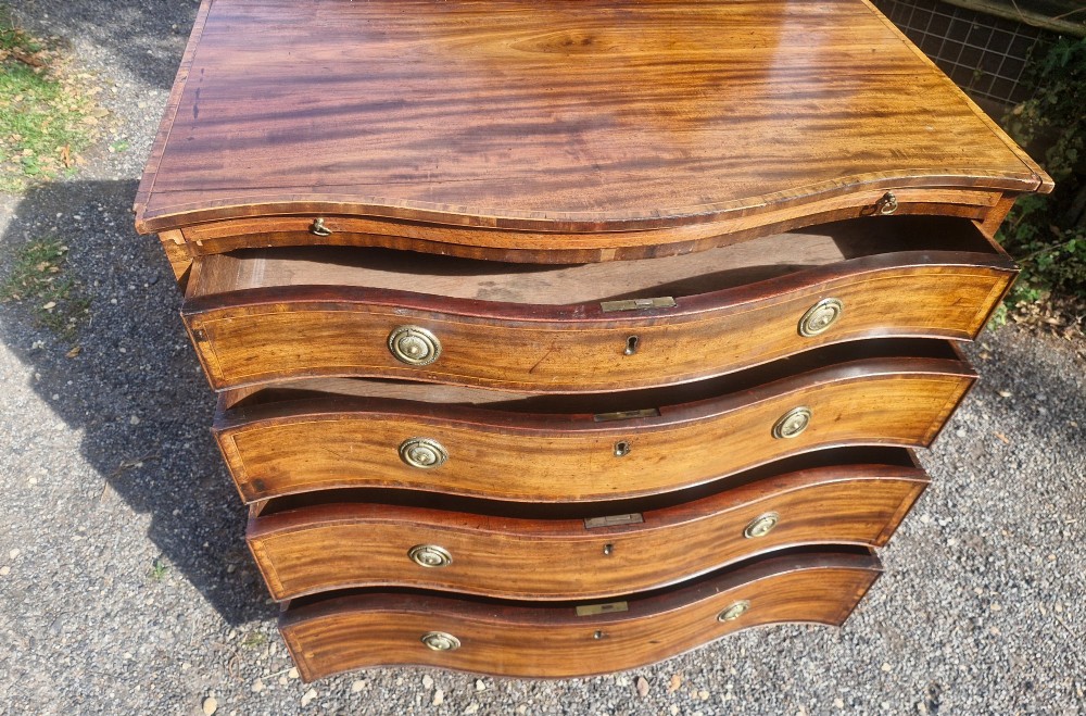 An antique mahogany chest of drawers featuring a serpentine curved front, four graduated drawers with circular brass pull handles, a pull-out brushing slide, and inlaid stringing detail. View IV