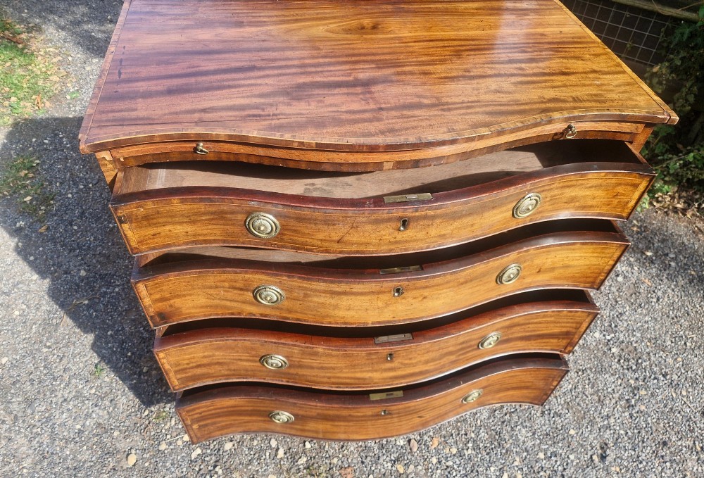 An antique mahogany chest of drawers featuring a serpentine curved front, four graduated drawers with circular brass pull handles, a pull-out brushing slide, and inlaid stringing detail. View V