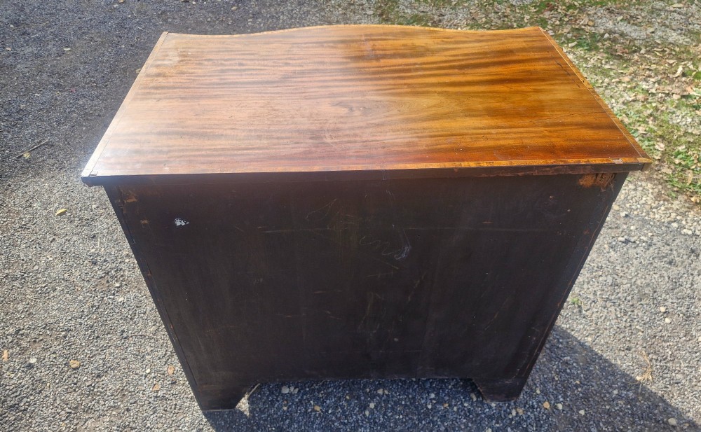 An antique mahogany chest of drawers featuring a serpentine curved front, four graduated drawers with circular brass pull handles, a pull-out brushing slide, and inlaid stringing detail. View VI
