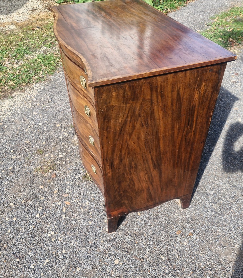 An antique mahogany chest of drawers featuring a serpentine curved front, four graduated drawers with circular brass pull handles, a pull-out brushing slide, and inlaid stringing detail. View VII