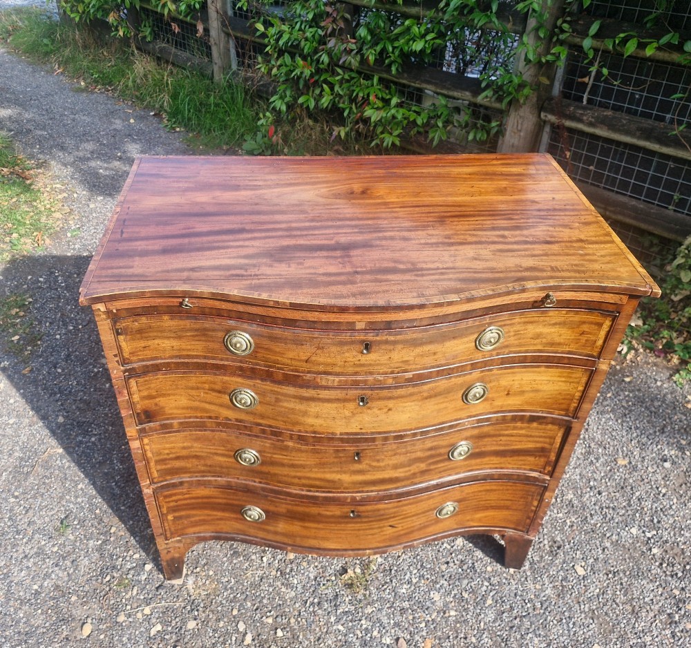 An antique mahogany chest of drawers featuring a serpentine curved front, four graduated drawers with circular brass pull handles, a pull-out brushing slide, and inlaid stringing detail. View VIII
