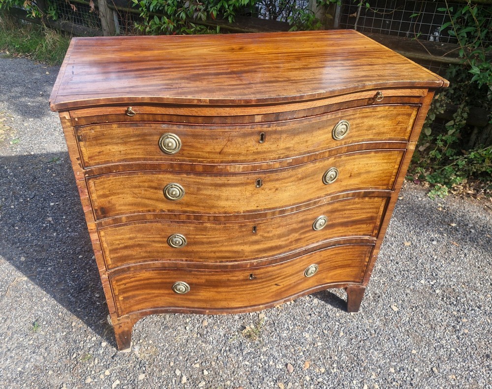 An antique mahogany chest of drawers featuring a serpentine curved front, four graduated drawers with circular brass pull handles, a pull-out brushing slide, and inlaid stringing detail. View IX