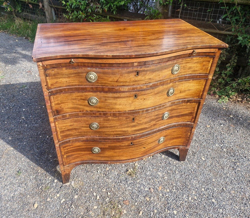 An antique mahogany chest of drawers featuring a serpentine curved front, four graduated drawers with circular brass pull handles, a pull-out brushing slide, and inlaid stringing detail. View X