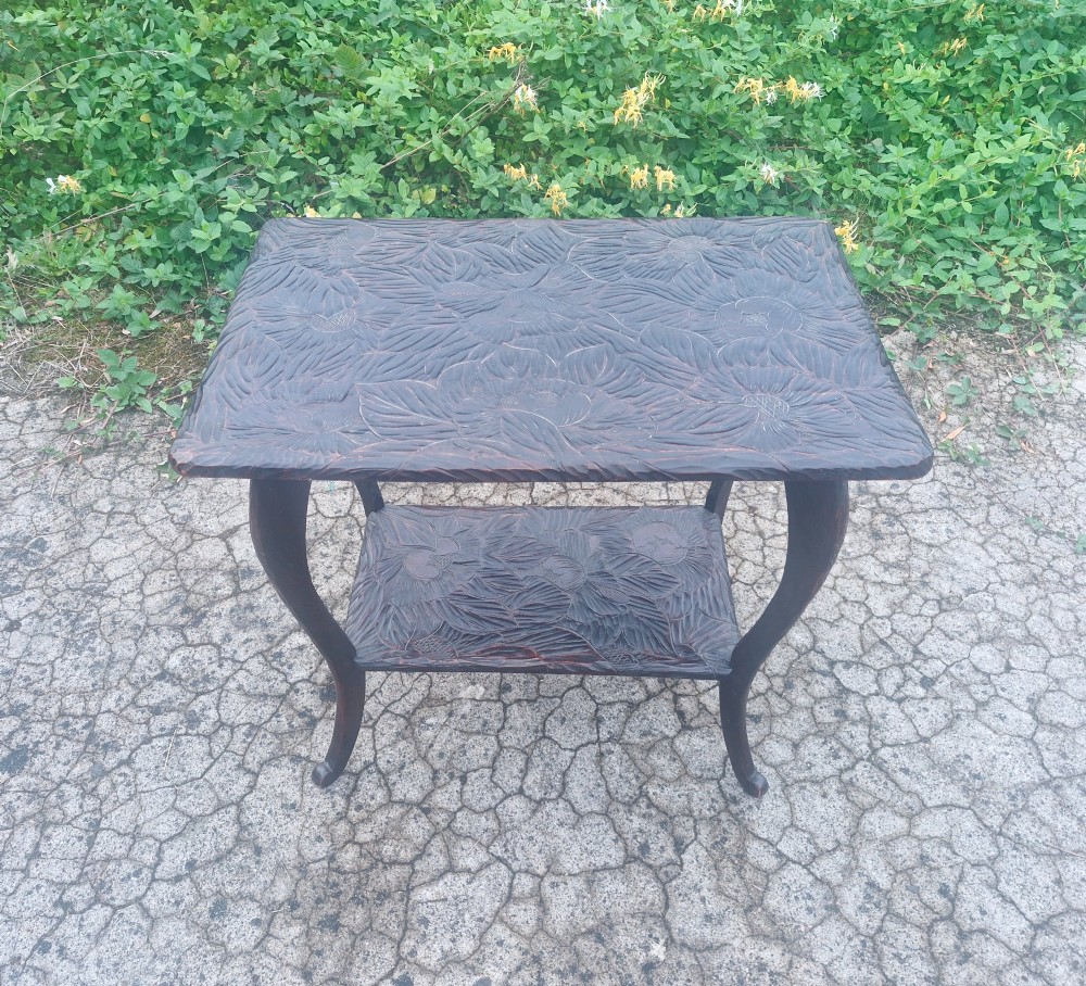 A dark brown two-tier wooden occasional table featuring intricate floral relief carvings across both flat surfaces and supported by four curved legs.