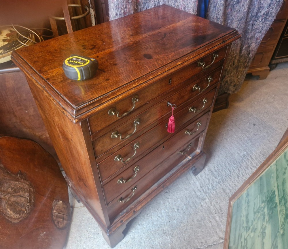 A Georgian oak chest of drawers featuring five graduated drawers with brass swan-neck handles and bracket feet. View V