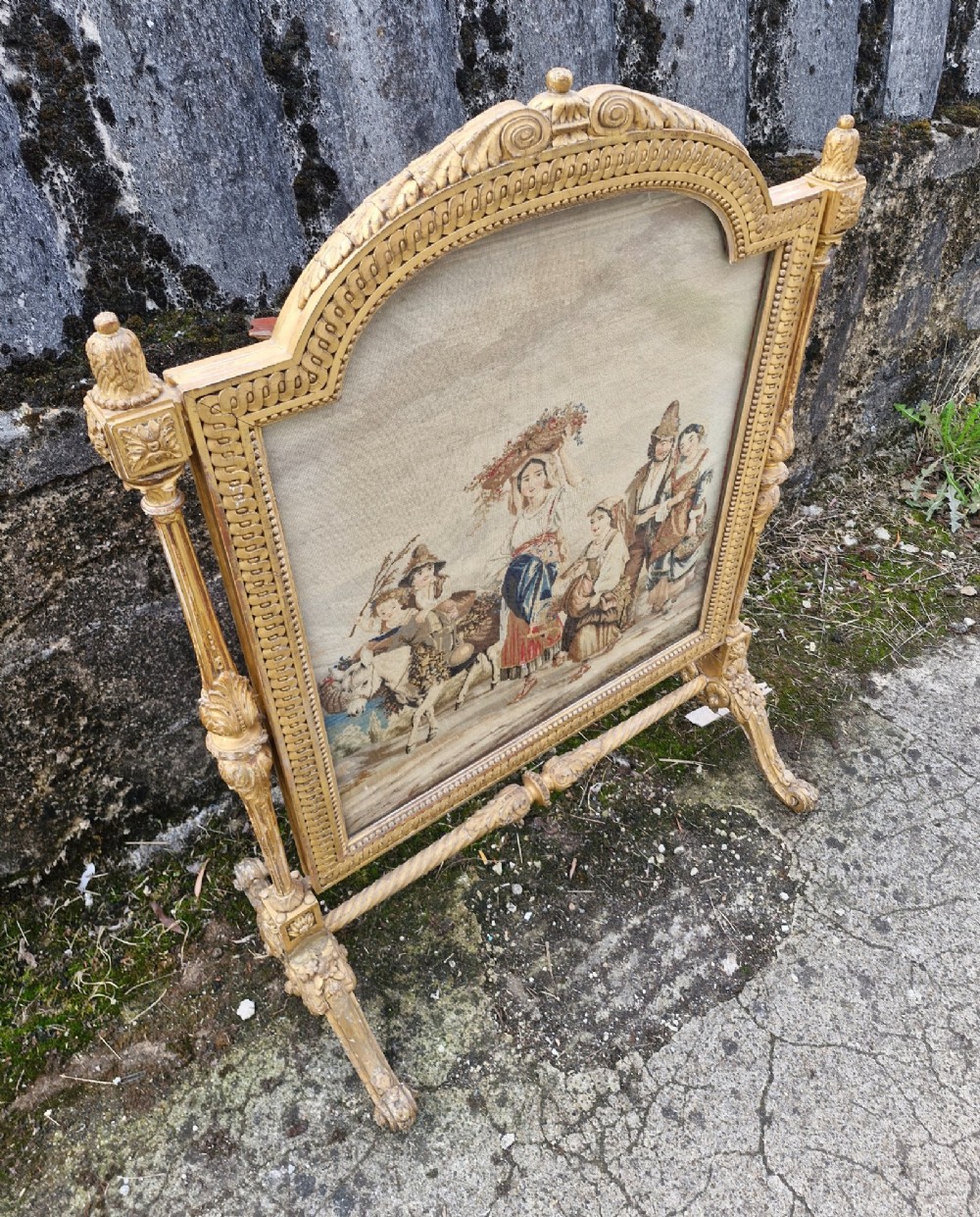 A Victorian carved giltwood fire screen featuring an arched frame, turned side columns, scroll feet, and a central tapestry panel depicting figures in traditional rural attire.