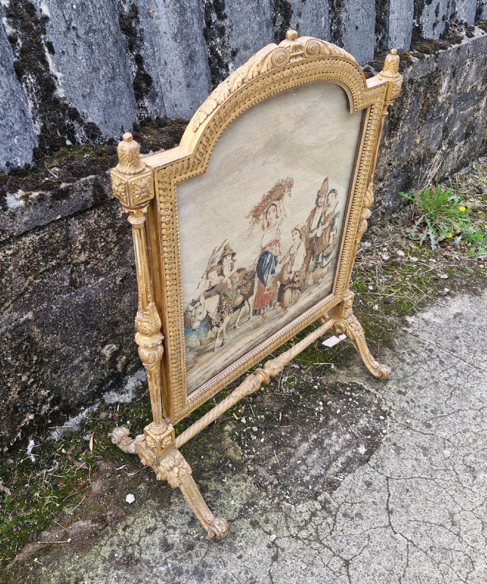 A Victorian carved giltwood fire screen featuring an arched frame, turned side columns, scroll feet, and a central tapestry panel depicting figures in traditional rural attire. View IV