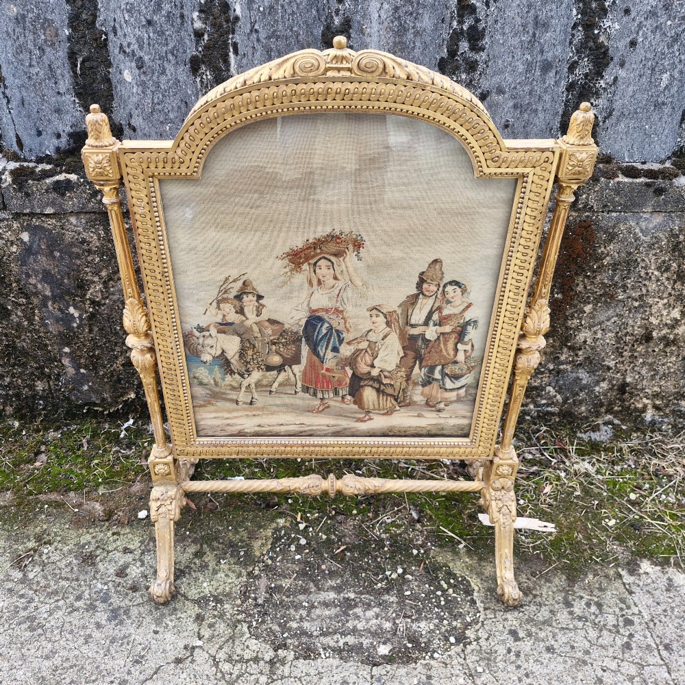 A Victorian carved giltwood fire screen featuring an arched frame, turned side columns, scroll feet, and a central tapestry panel depicting figures in traditional rural attire. View VI