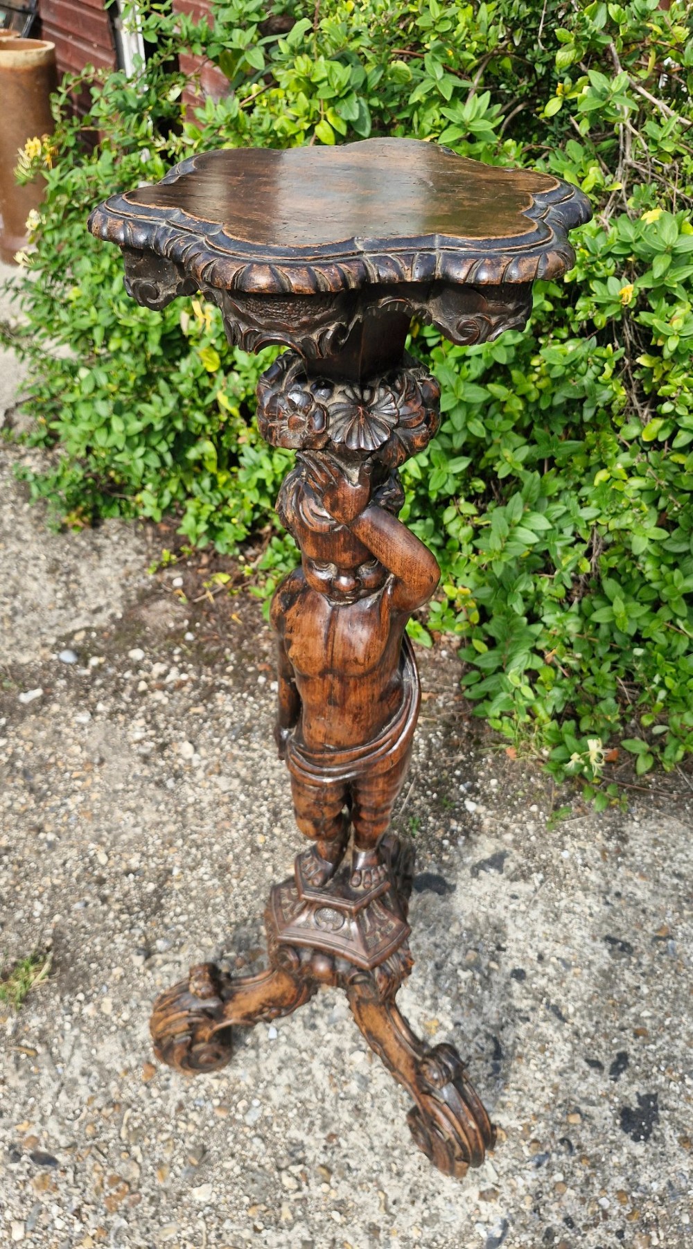 A 19th-century Italian dark carved wood pedestal stand with a putto figure support, scalloped top, and scroll tripod feet.