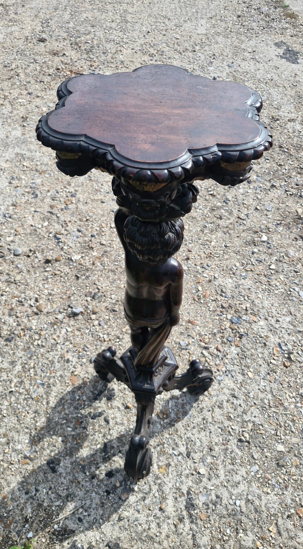 A carved dark wood pedestal stand featuring a central putto figure supporting a shaped top with gilt decorative accents on a tripod scroll base. View II