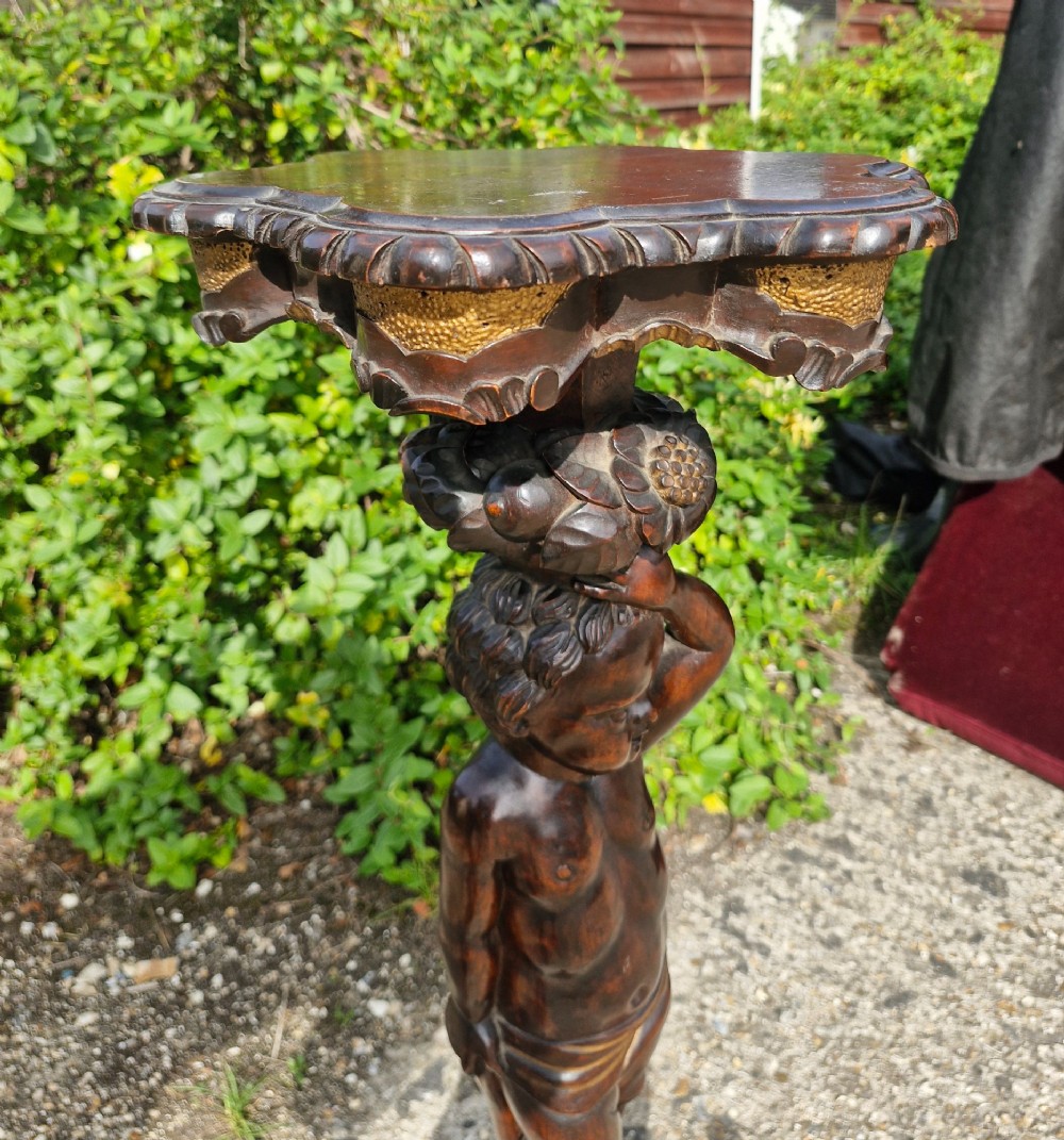 A carved dark wood pedestal stand featuring a central putto figure supporting a shaped top with gilt decorative accents on a tripod scroll base. View III