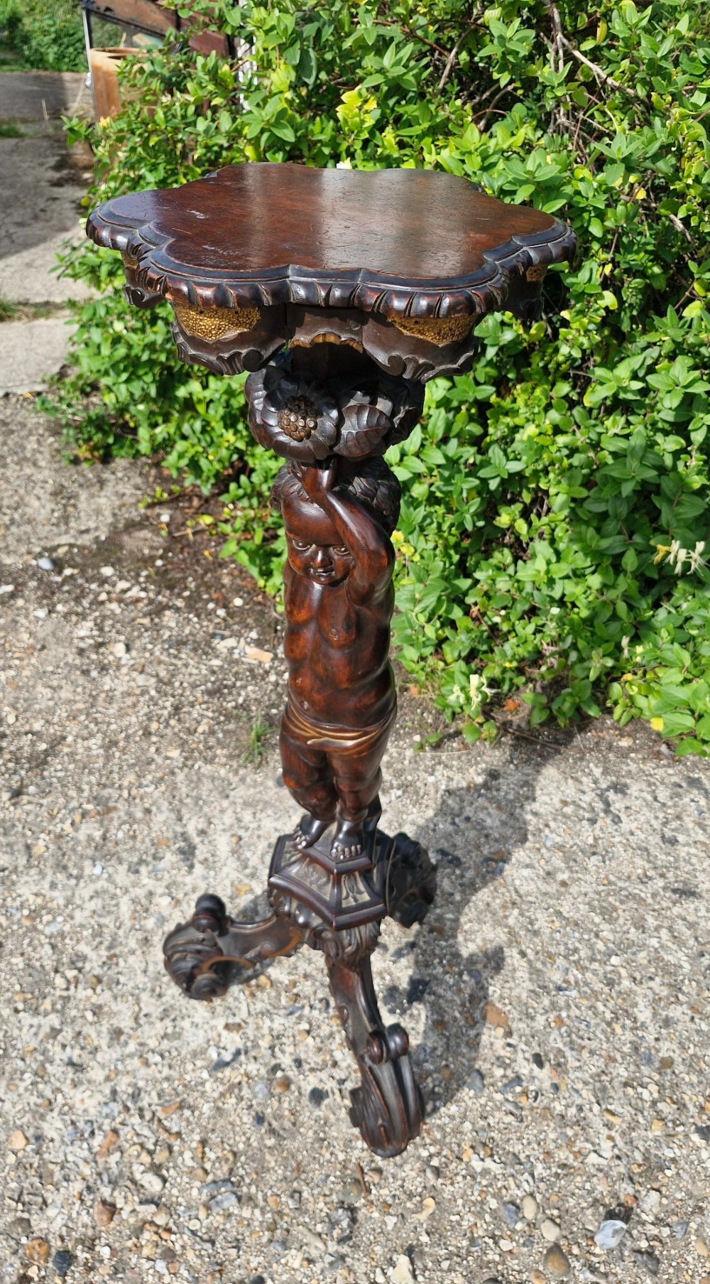 A carved dark wood pedestal stand featuring a central putto figure supporting a shaped top with gilt decorative accents on a tripod scroll base. View IV