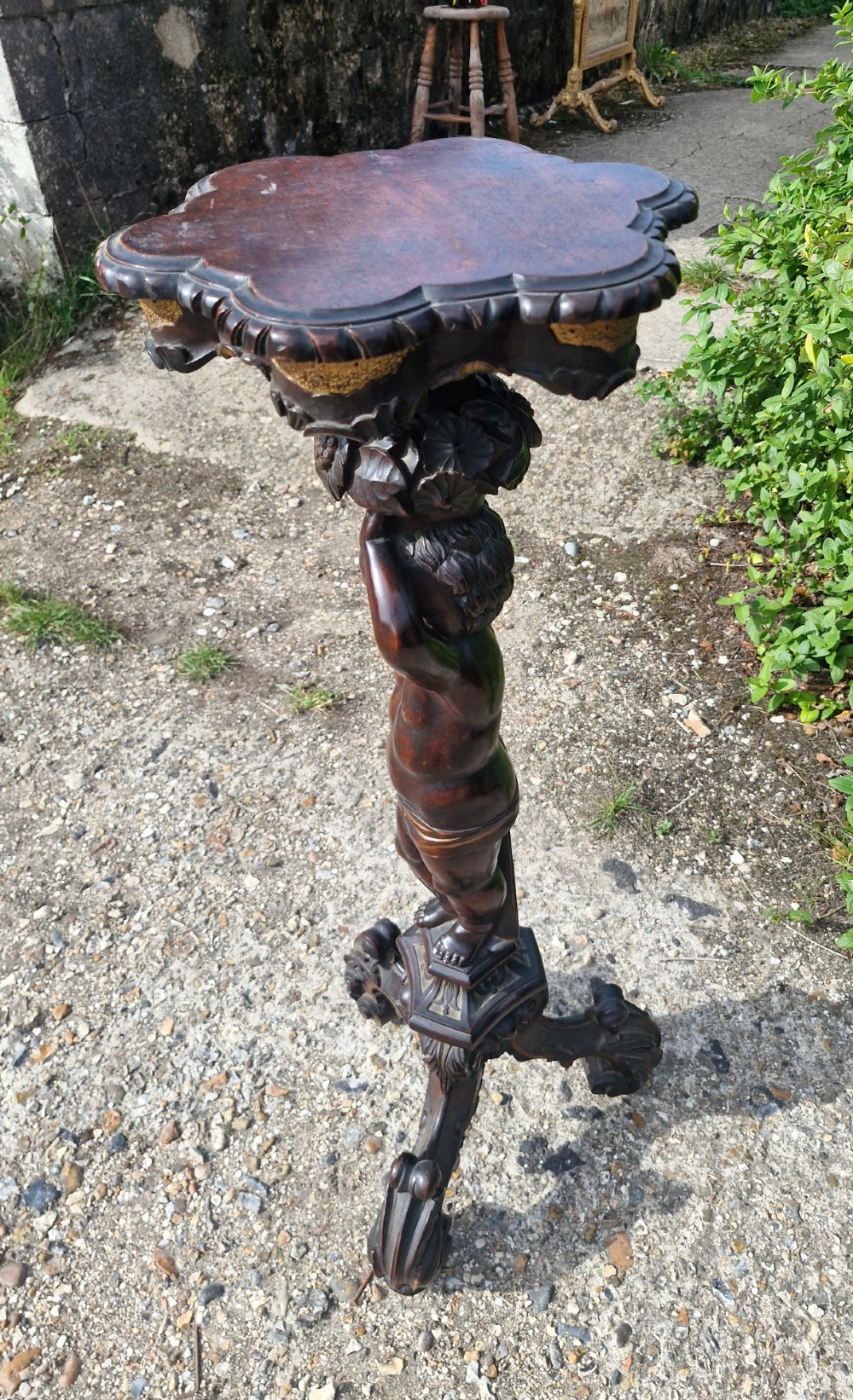 A carved dark wood pedestal stand featuring a central putto figure supporting a shaped top with gilt decorative accents on a tripod scroll base. View V