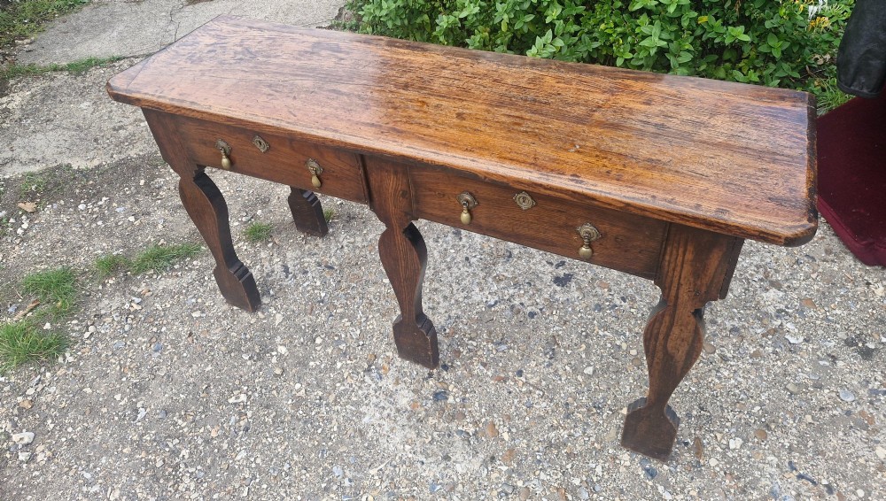 An early 19th-century country oak serving table featuring a long rectangular top, two drawers with brass teardrop handles, and shaped flat-profile legs. View X