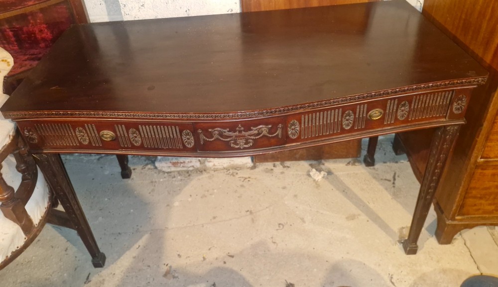 An Edwardian dark mahogany serving table featuring a shaped bow front, carved swags and fluted frieze decoration, two drawers, and square tapered legs.