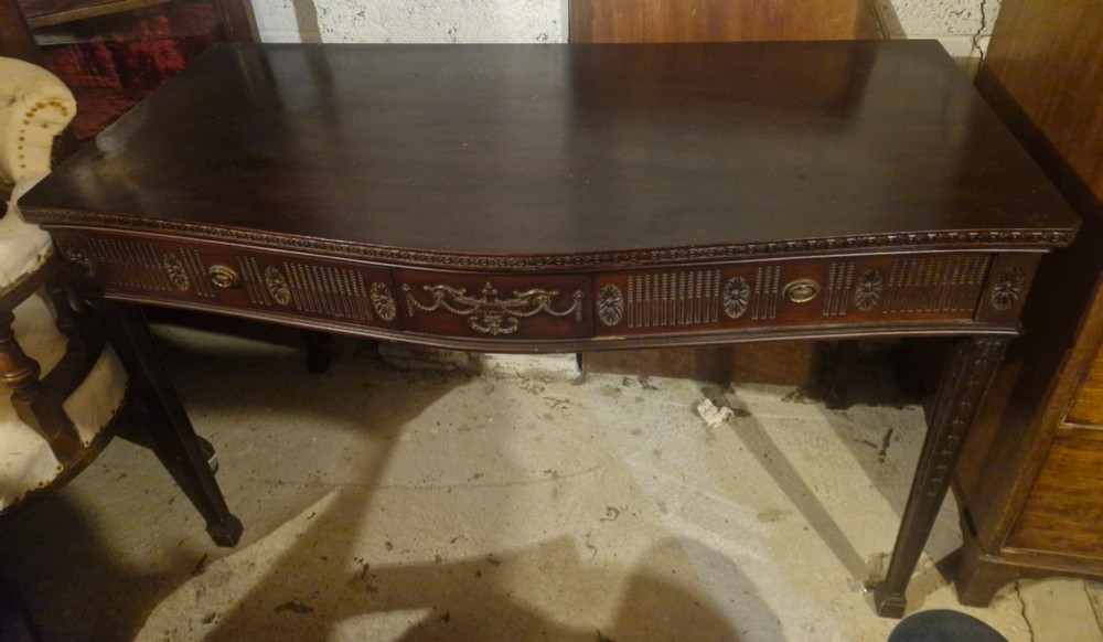 An Edwardian dark mahogany serving table featuring a shaped bow front, carved swags and fluted frieze decoration, two drawers, and square tapered legs. View II