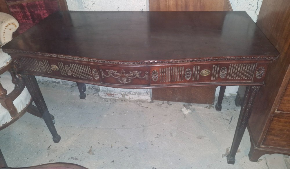 An Edwardian dark mahogany serving table featuring a shaped bow front, carved swags and fluted frieze decoration, two drawers, and square tapered legs. View V