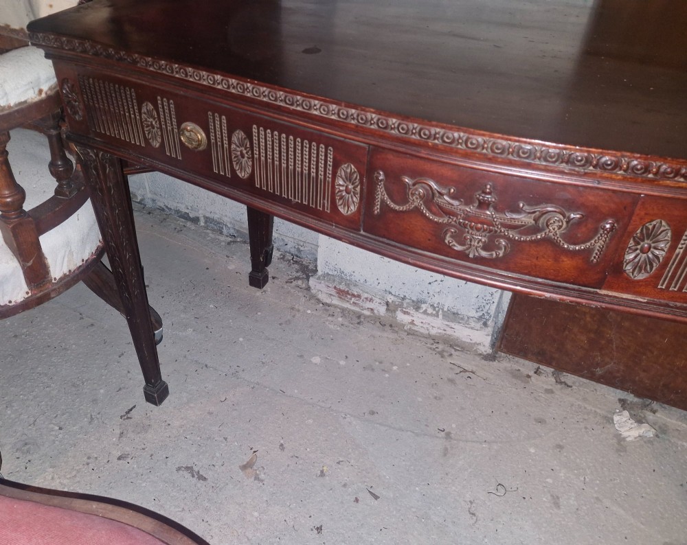 An Edwardian dark mahogany serving table featuring a shaped bow front, carved swags and fluted frieze decoration, two drawers, and square tapered legs. View VI