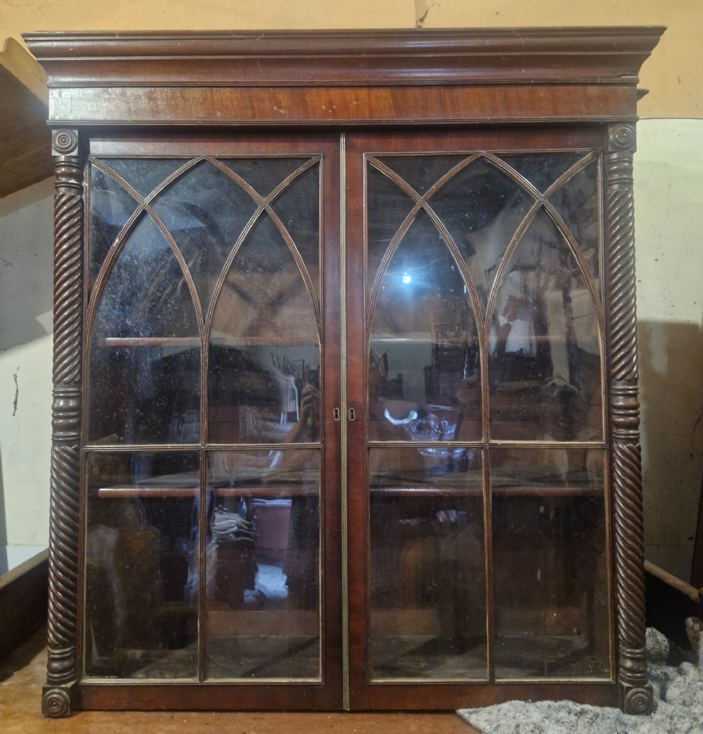 A Regency mahogany wall cabinet featuring two Gothic astragal glazed doors, spiral-turned side columns, and carved roundel corner details.