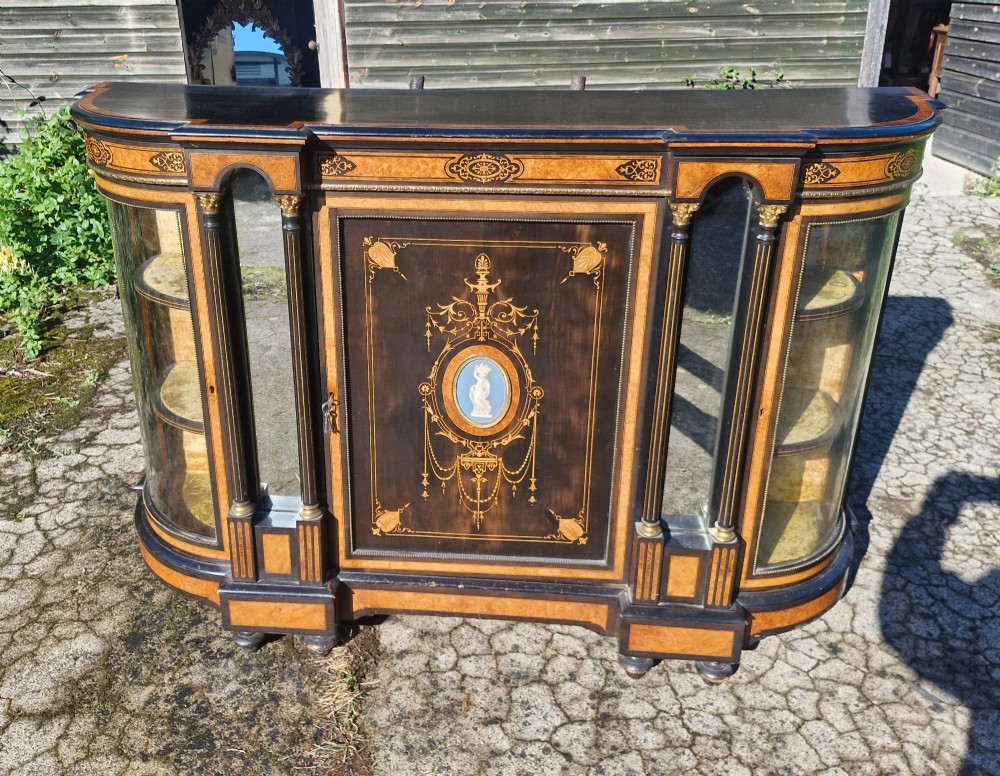 A Victorian ebonised side cabinet with intricate wood marquetry, curved glass display sides, gilt column capitals, and a central blue porcelain oval medallion.