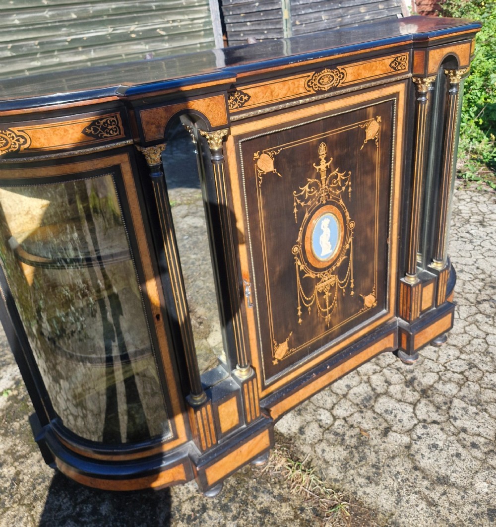 A Victorian ebonised side cabinet with intricate wood marquetry, curved glass display sides, gilt column capitals, and a central blue porcelain oval medallion. View III