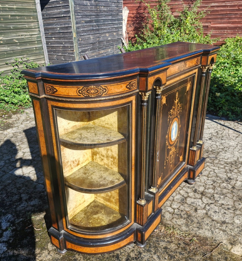A Victorian ebonised side cabinet with intricate wood marquetry, curved glass display sides, gilt column capitals, and a central blue porcelain oval medallion. View IV