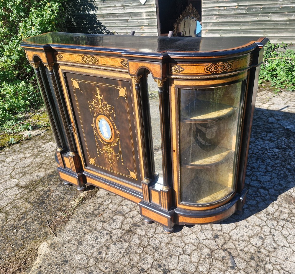 A Victorian ebonised side cabinet with intricate wood marquetry, curved glass display sides, gilt column capitals, and a central blue porcelain oval medallion. View V