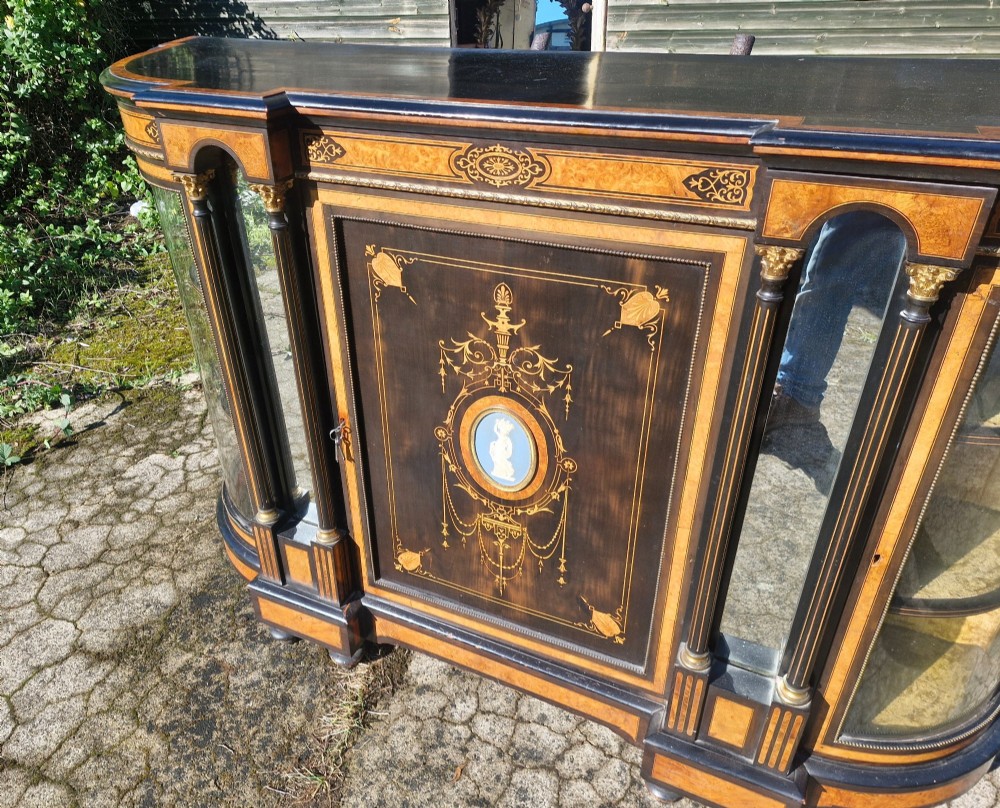 A Victorian ebonised side cabinet with intricate wood marquetry, curved glass display sides, gilt column capitals, and a central blue porcelain oval medallion. View VI