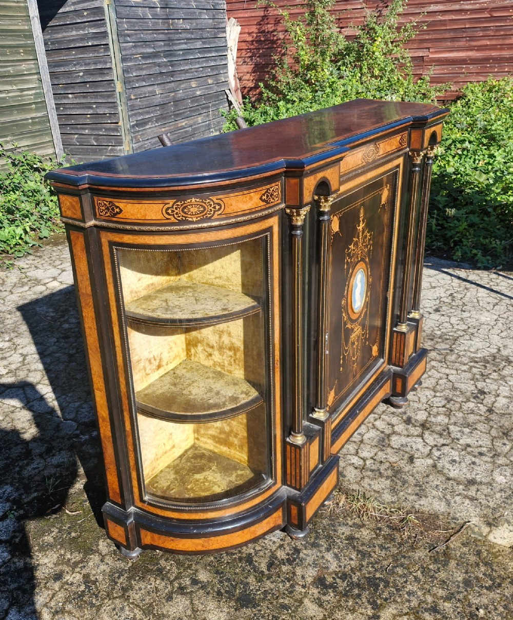 A Victorian ebonised side cabinet with intricate wood marquetry, curved glass display sides, gilt column capitals, and a central blue porcelain oval medallion. View VII