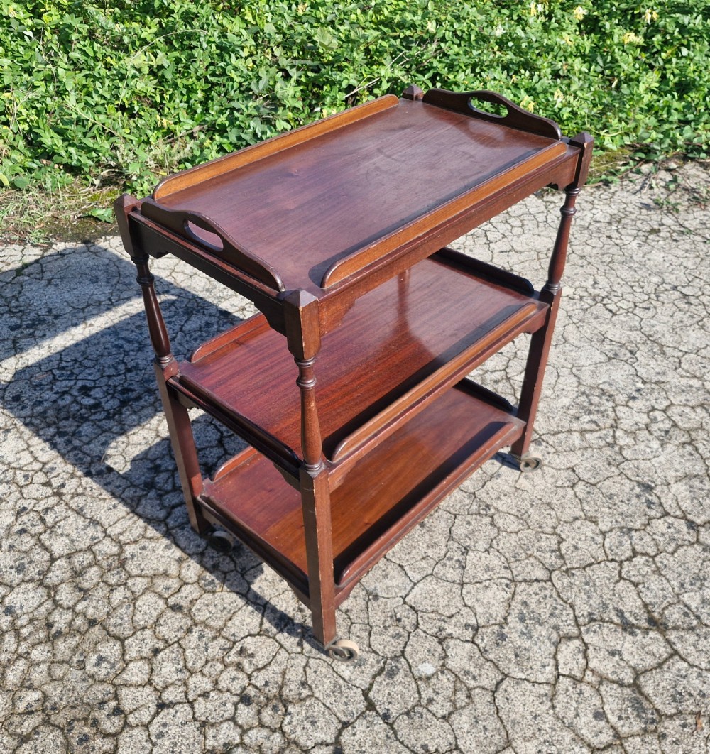 A three-tier dark mahogany wooden tea trolley featuring turned corner posts, castor wheels, and a top removable tray with cut-out handles.