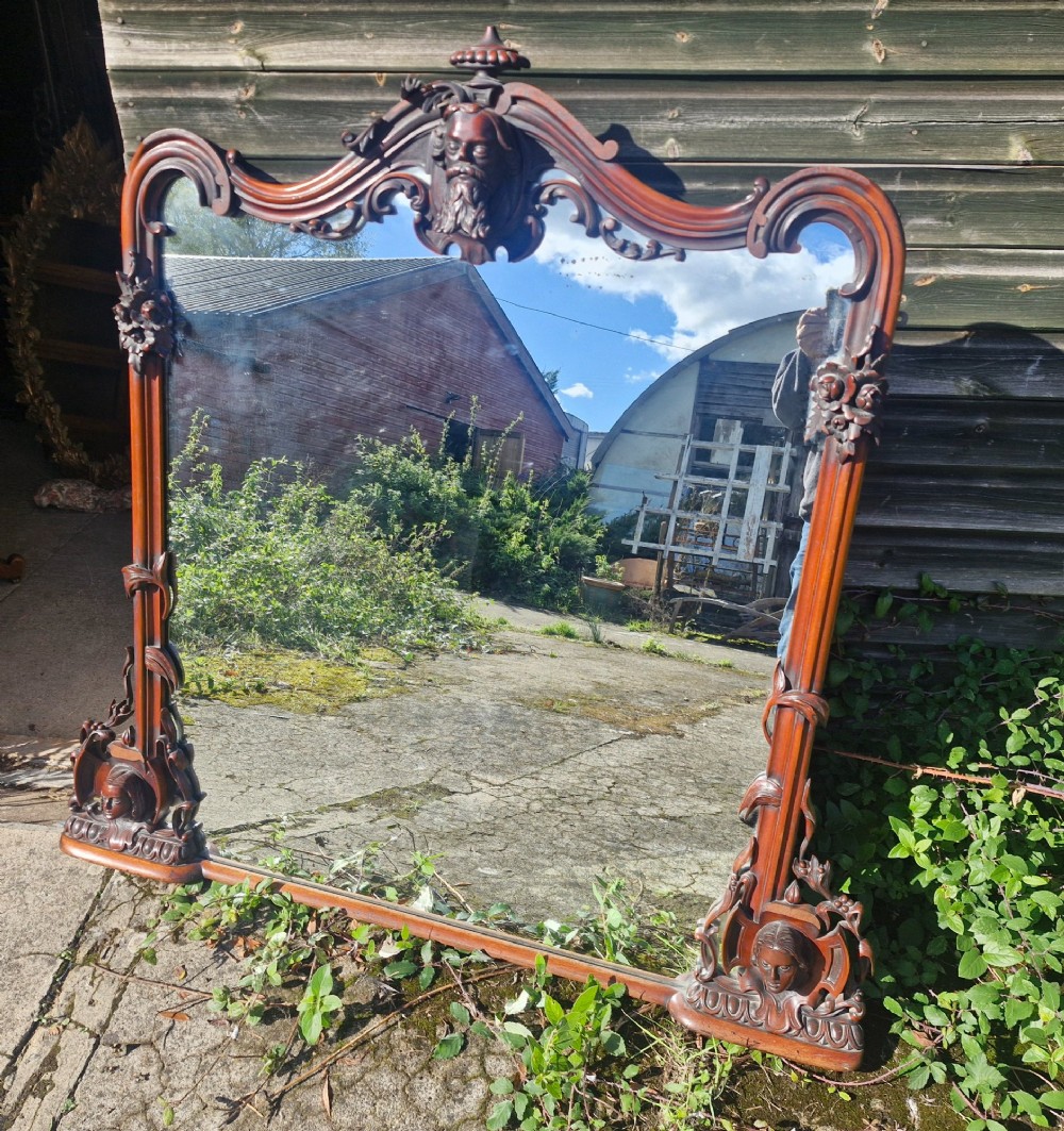 An antique Victorian carved mahogany overmantel mirror featuring an arched frame decorated with carved human faces, scrollwork, and foliate details.