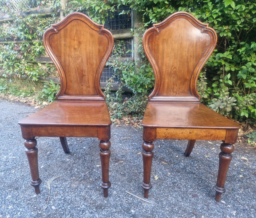 A pair of Victorian timber hall chairs featuring shaped back panels, solid wooden seats, and turned front legs.