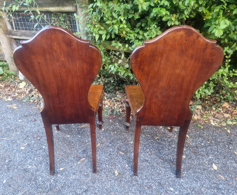 A pair of Victorian timber hall chairs featuring shaped back panels, solid wooden seats, and turned front legs. View V