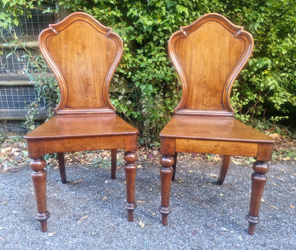 A pair of Victorian timber hall chairs featuring shaped back panels, solid wooden seats, and turned front legs. View VI