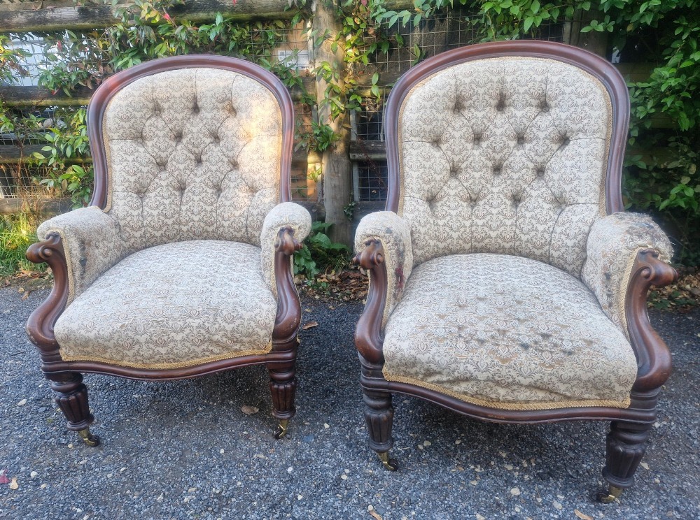 A pair of Victorian mahogany library armchairs featuring deep button-back patterned upholstery, carved scroll arms, and turned legs with brass castors. View VI