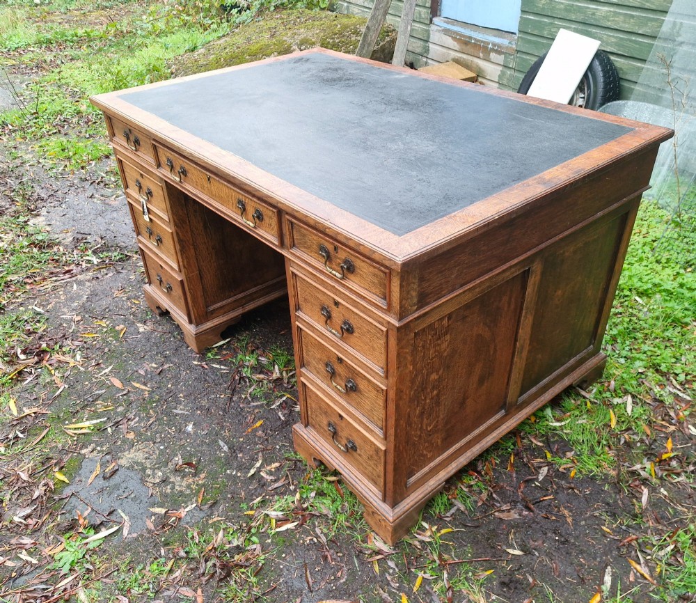 An antique oak pedestal writing desk featuring a black inset leather writing surface, multiple drawers with brass drop handles, and panelled sides.