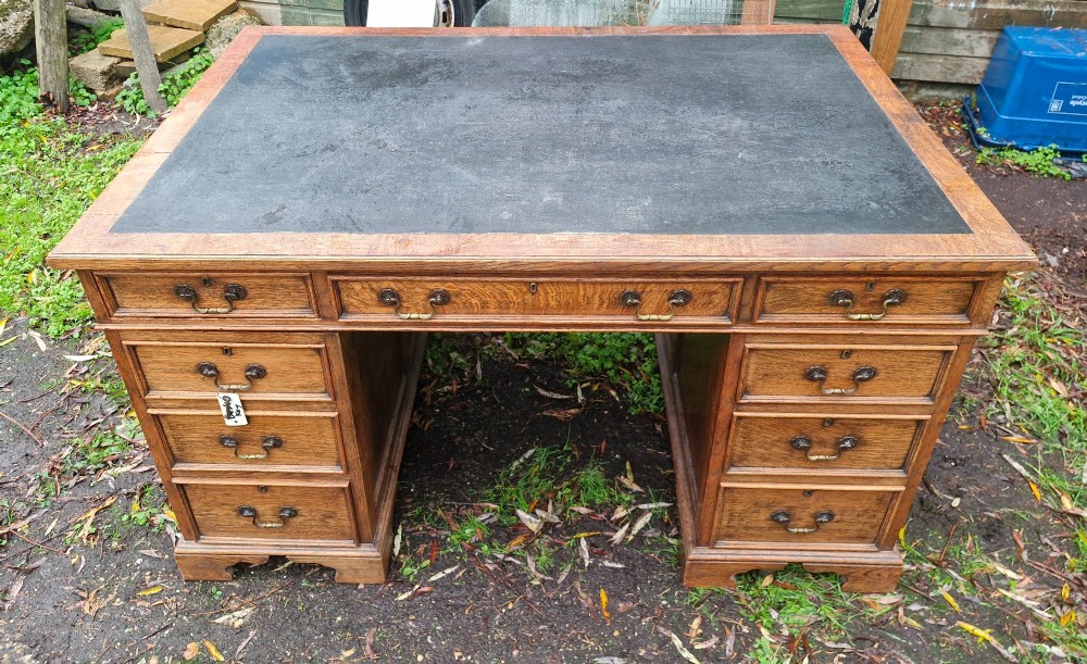 An antique oak pedestal writing desk featuring a black inset leather writing surface, multiple drawers with brass drop handles, and panelled sides. View II