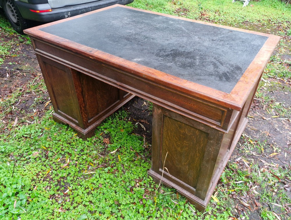 An antique oak pedestal writing desk featuring a black inset leather writing surface, multiple drawers with brass drop handles, and panelled sides. View III